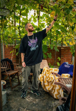 Load image into Gallery viewer, man wearing black oversized Plant A Seed floral graphic t-shirt with green oversized green roboflower cargos and puma rs-x shoes in a green house