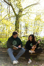 Load image into Gallery viewer, man wearing oversized green tree of life hoodie while sketching, woman wearing black oversized tree of life t-shirt while playing a banjo
