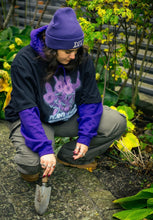 Load image into Gallery viewer, woman wearing layered, oversized black plant a seed t-shirt with green roboflower cargos and purple DFKT logo beanie while diggin in garden