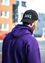 Load image into Gallery viewer, man wearing black DFKT logo dad hat cap backwards with purple hoodie