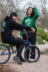 Woman wearing green oversized roboflower graphic sweater, man wearing black fly outside the jar butterfly graphic hoodie