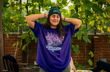 Load image into Gallery viewer, woman wearing oversized purple Plant A Seed floral eye graphic t-shirt with green DFKT logo beanie