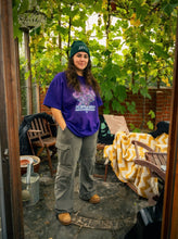 Load image into Gallery viewer, woman wearing purple oversized plant a seed eye graphic t-shirt with green roboflower cargos and green DFKT logo beanie