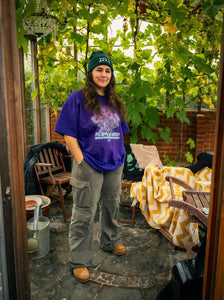woman wearing purple oversized plant a seed eye graphic t-shirt with green roboflower cargos and green DFKT logo beanie