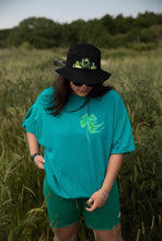 Load image into Gallery viewer, woman wearing oversized green no luck t-shirt with bucket hat and green shorts in a field of wheat